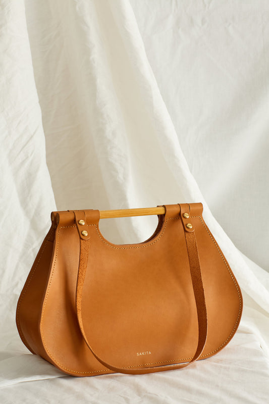 Angled view of tan leather tote bag with curved shape and top wooden handles, photographed in soft light