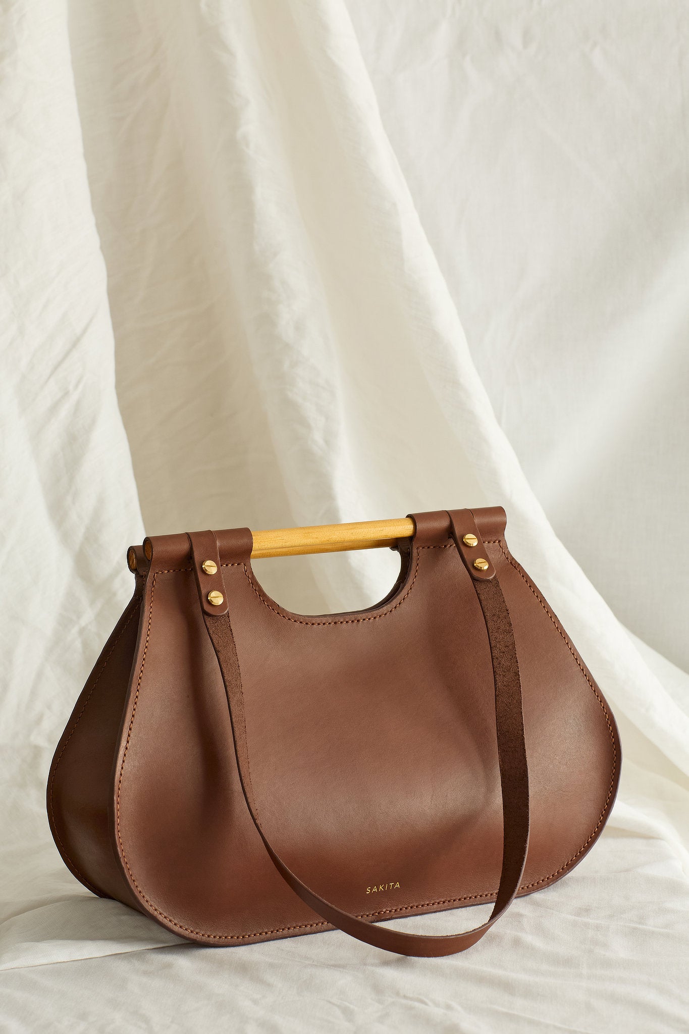Angled view of brown leather tote bag with curved shape and top wooden handles, photographed in soft light