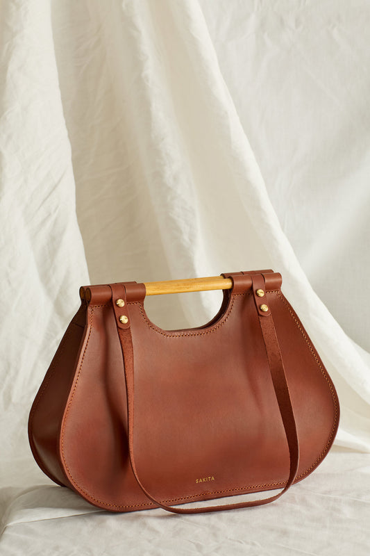 Angled view of maroon leather tote bag with curved shape and top wooden handles, photographed in soft light