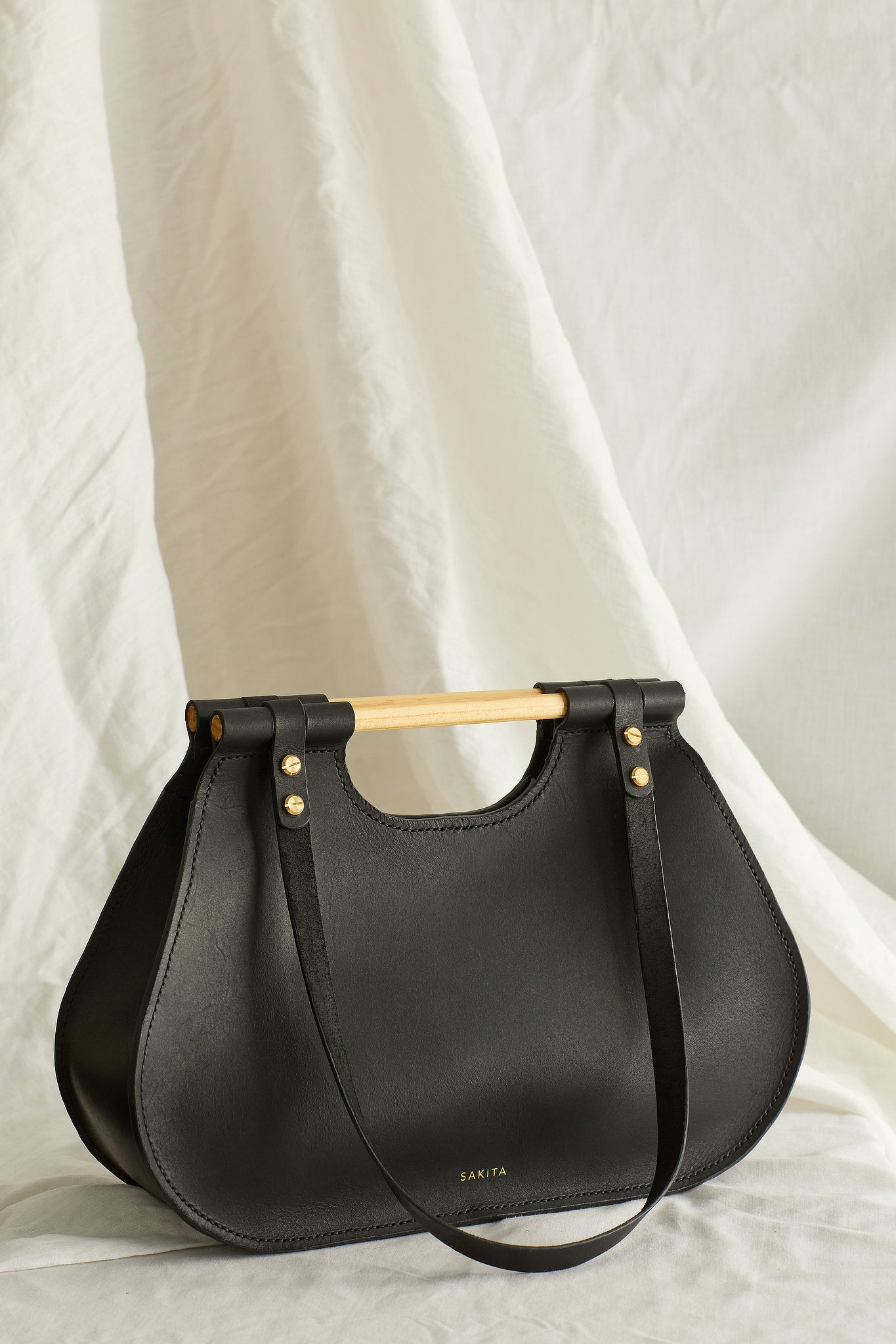 Angled view of black leather tote bag with curved shape and top wooden handles, photographed in soft light