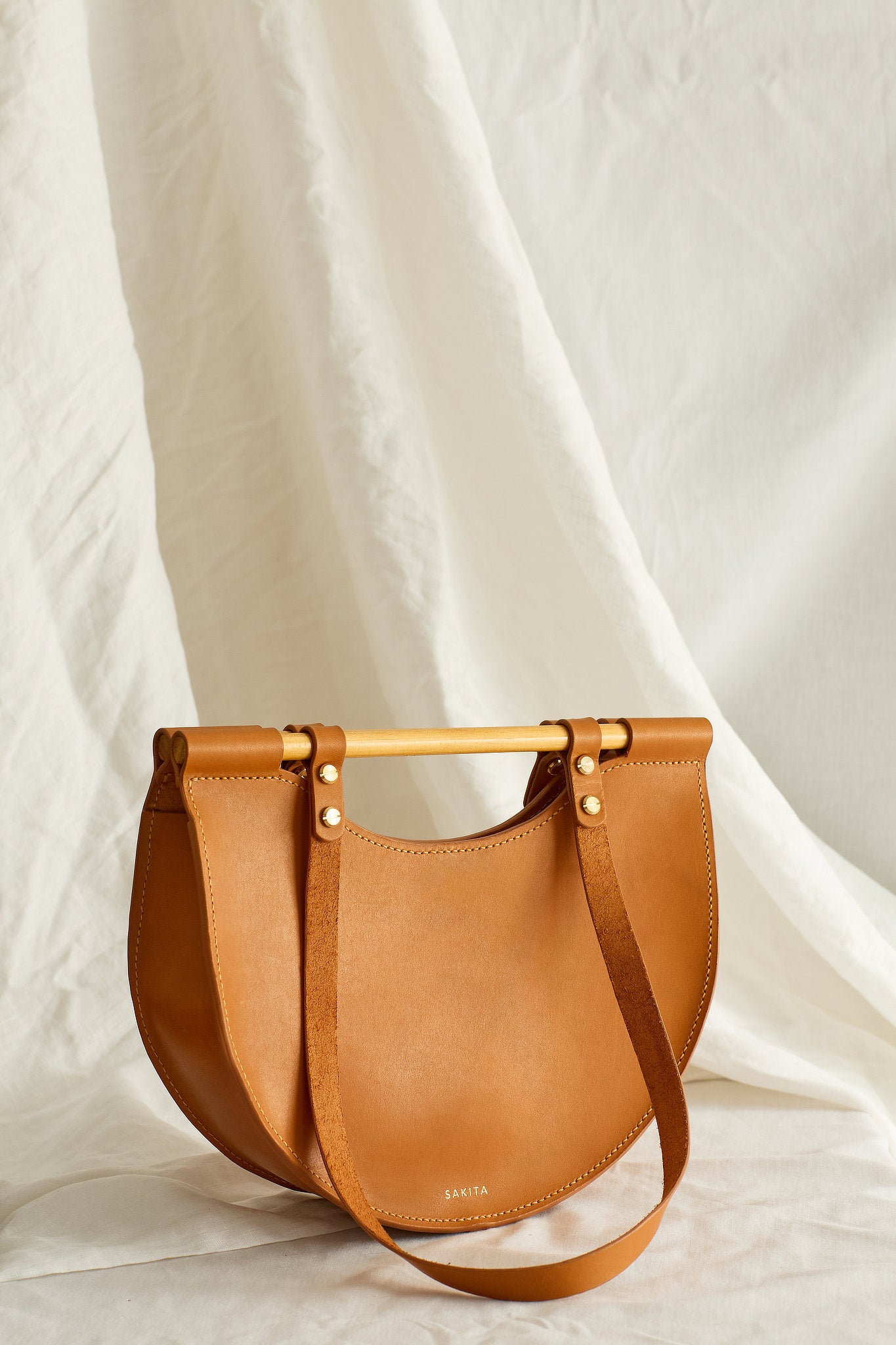 Angled view of tan leather tote bag with half moon shape and top wooden handles, photographed in soft light