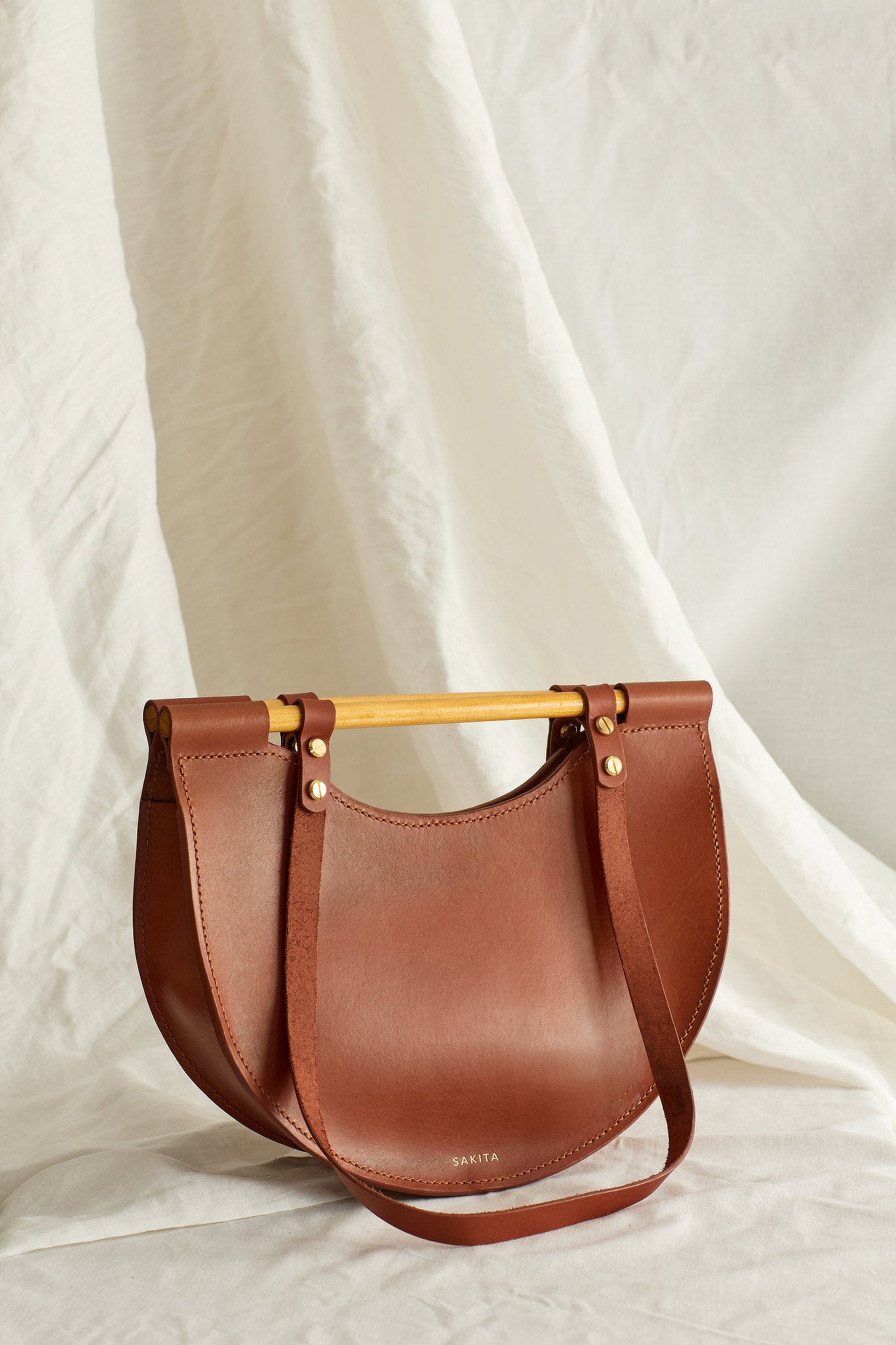 Angled view of maroon leather tote bag with half moon shape and top wooden handles, photographed in soft light
