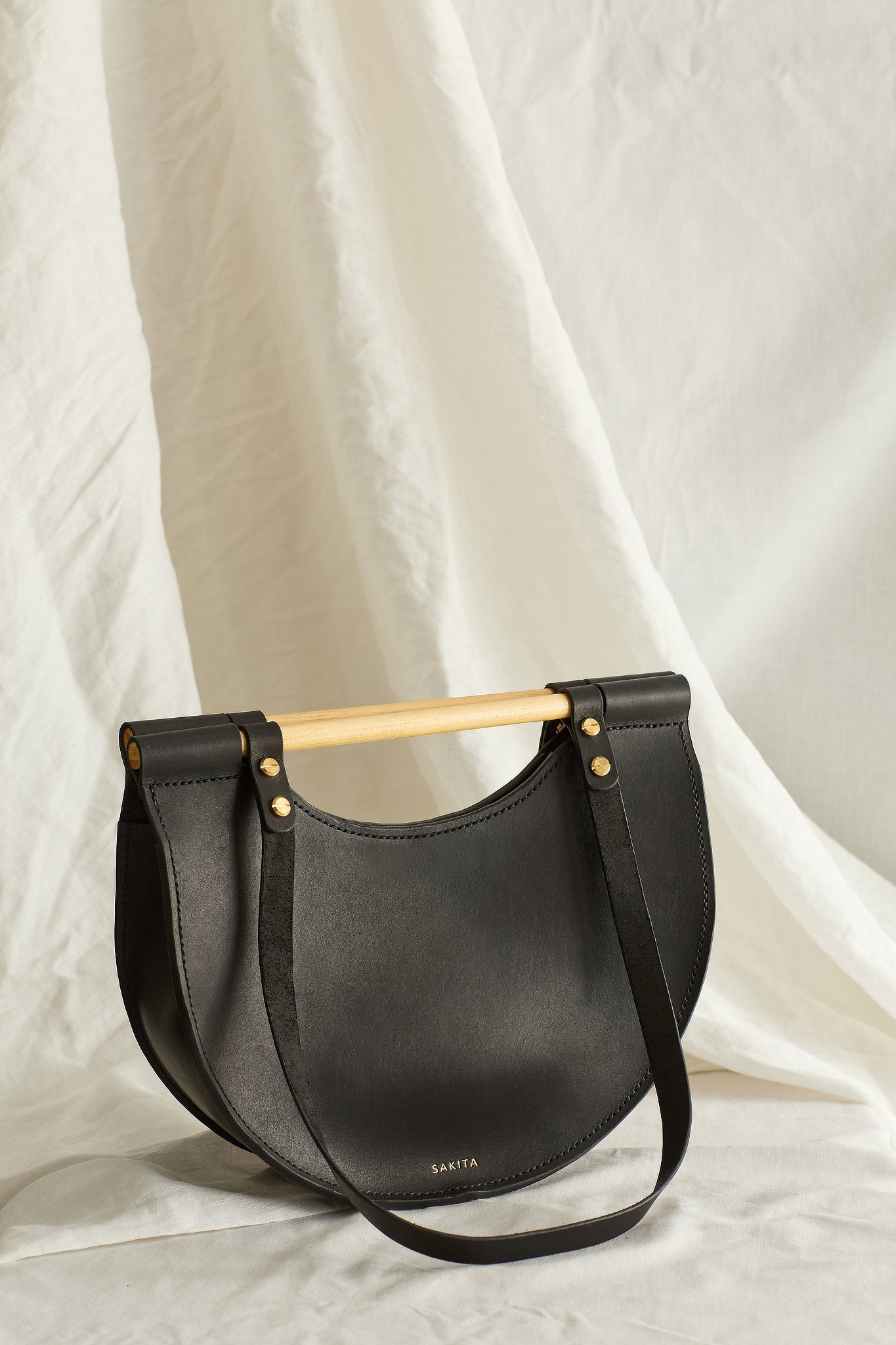 Angled view of black leather tote bag with half moon shape and top wooden handles, photographed in soft light