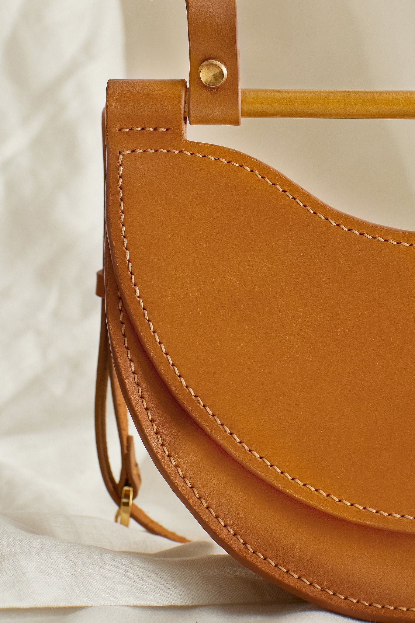 Close-up of small tan leather half moon bag showing hand-stitching, brass hardware, and wooden dowel detail