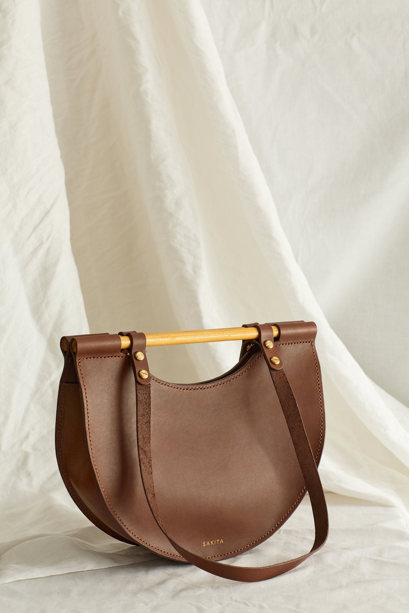 Angled view of brown leather tote bag with half moon shape and top wooden handles, photographed in soft light